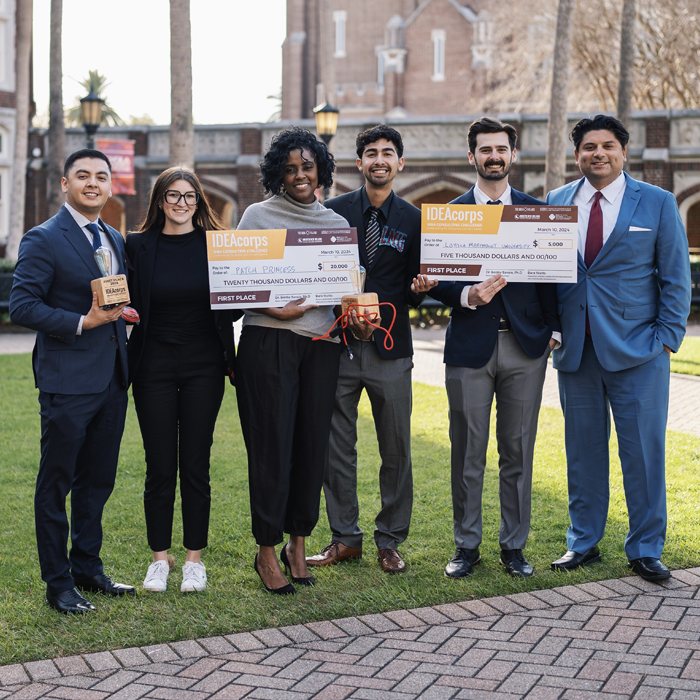 The IDEACorps MBA Consulting Challenge 2024 Team with their faculty advisor and client holding up two large checks.